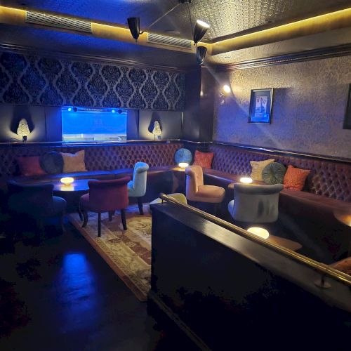 A cozy lounge with velvet booths, upholstered chairs, warm lamps and blue accent lighting, plus framed art on textured walls.