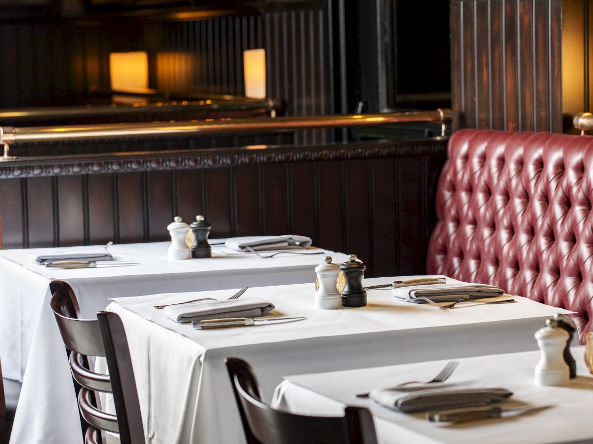 The image shows a restaurant with white tablecloths, dark wooden chairs, and a red upholstered booth. Tables are set with napkins and cutlery.