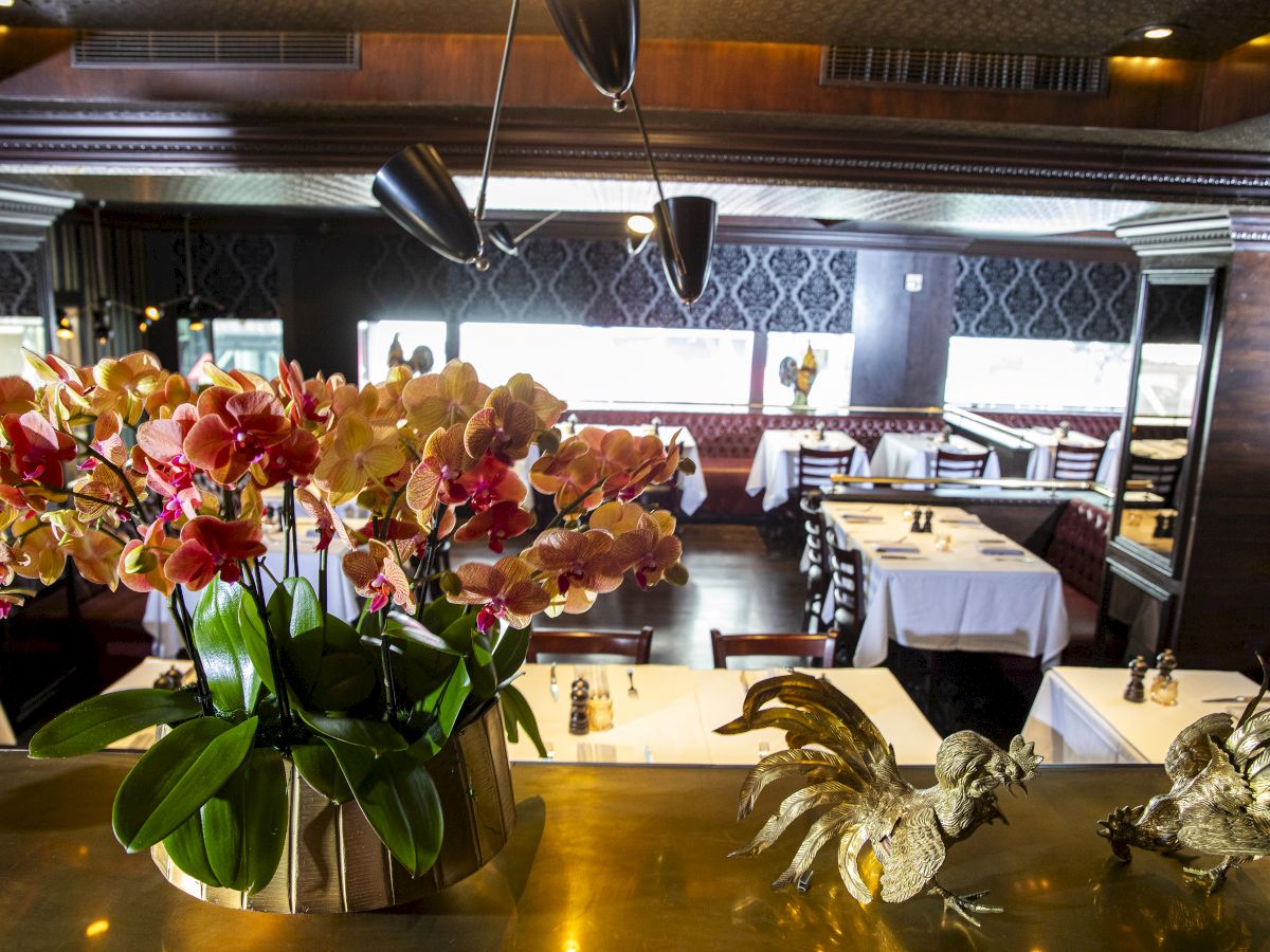 The image shows an elegant restaurant interior with decorated tables, colorful flowers in a vase, and rooster statues on a counter.