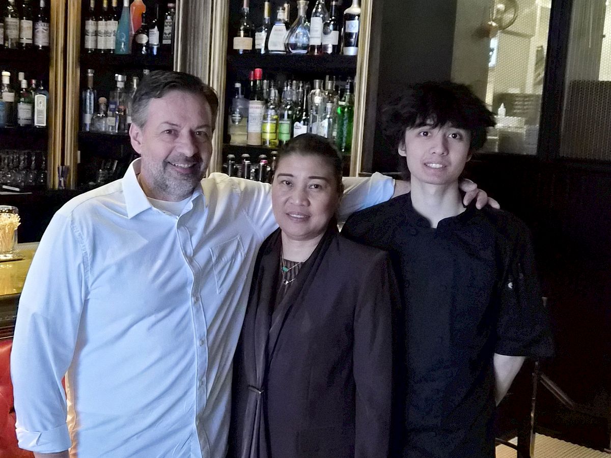 Three people are posing together at a bar, smiling in front of shelves packed with bottles of liquor.