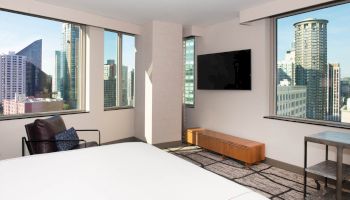 The image shows a modern hotel room with large windows offering a cityscape view, a TV, a chair, and a bed.