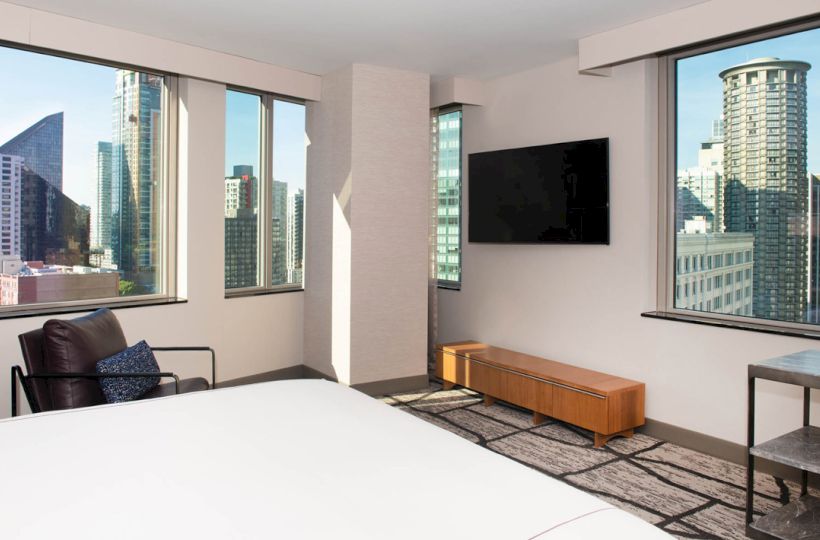 The image shows a modern hotel room with large windows offering a cityscape view, a TV, a chair, and a bed.