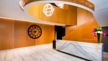 The image shows a modern lobby with a marble reception desk, a large wall clock, spiral wooden architecture, and decorative light fixtures.