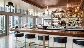A modern bar with a fully stocked shelf, counter seating, and overhead pendant lights, featuring large windows with city views in the background.