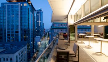 The image shows a modern high-rise cityscape at dusk with a stylish outdoor terrace featuring tables, chairs, and an open indoor area.
