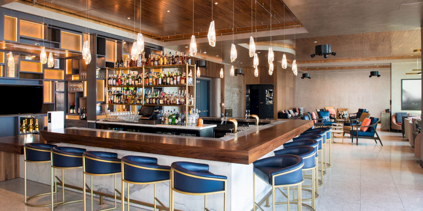 The image depicts a modern bar with a wooden counter, blue and gold barstools, hanging lights, and a fully stocked bar.