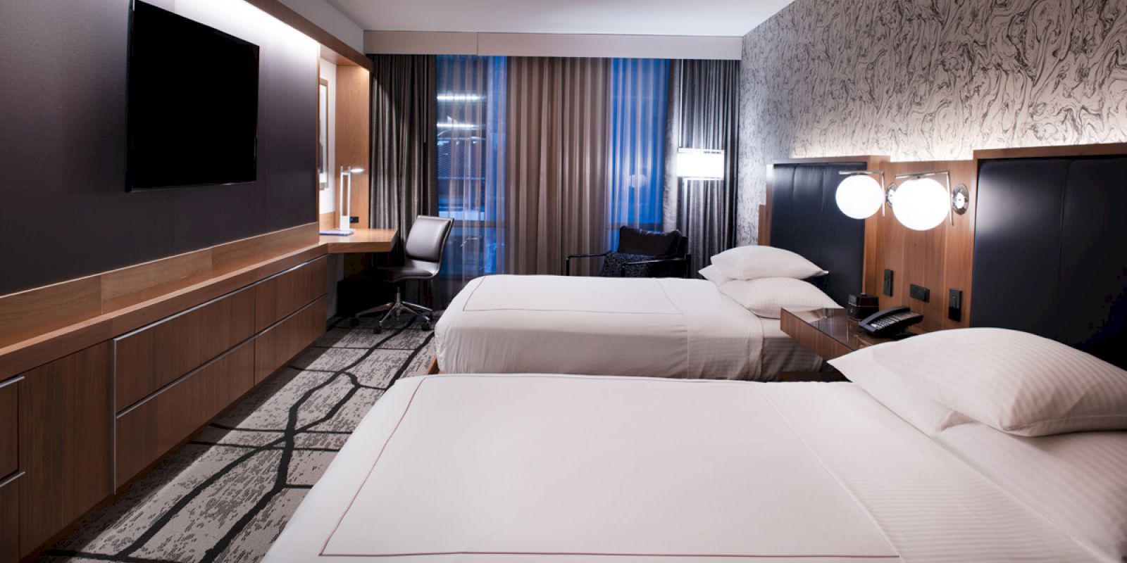 A modern hotel room with two neatly made beds, a large TV, a desk with a chair, and contemporary decor, featuring a patterned wall.