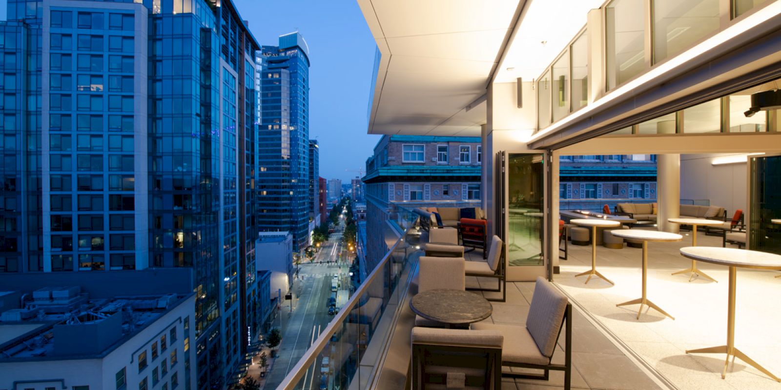 A modern balcony with seating, overlooking a city street lined with tall buildings, illuminated at dusk.