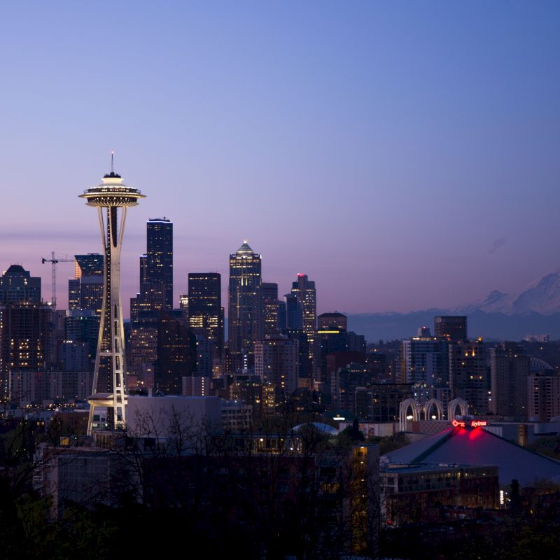 A city skyline at dusk featuring a prominent tower, skyscrapers, and a mountainous backdrop in the distance under a purple-blue sky.
