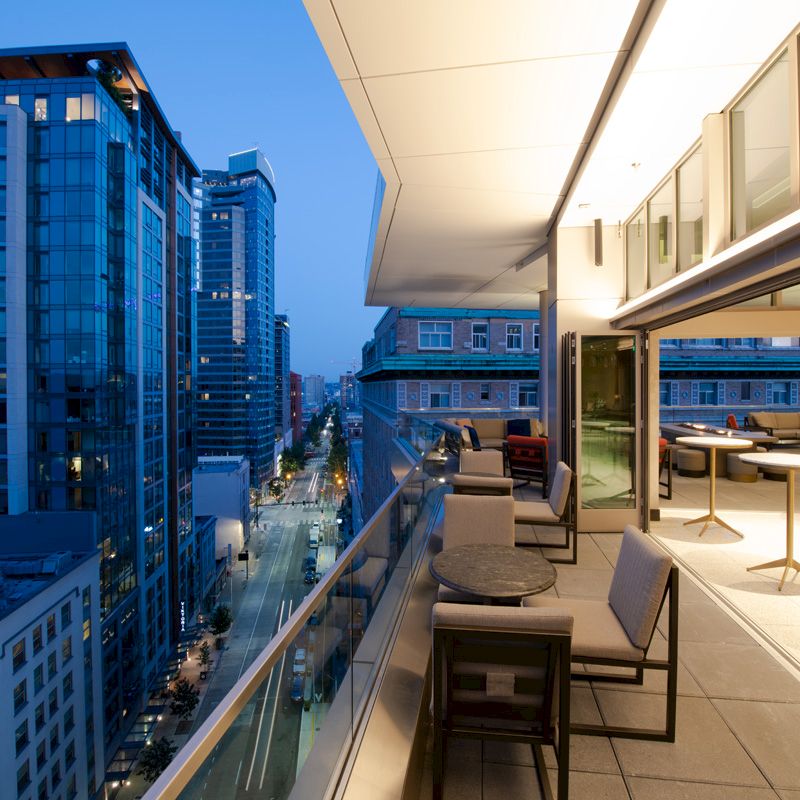 A modern cityscape at dusk featuring a balcony with seating and tables, overlooking a street lined with tall buildings illuminated by lights.