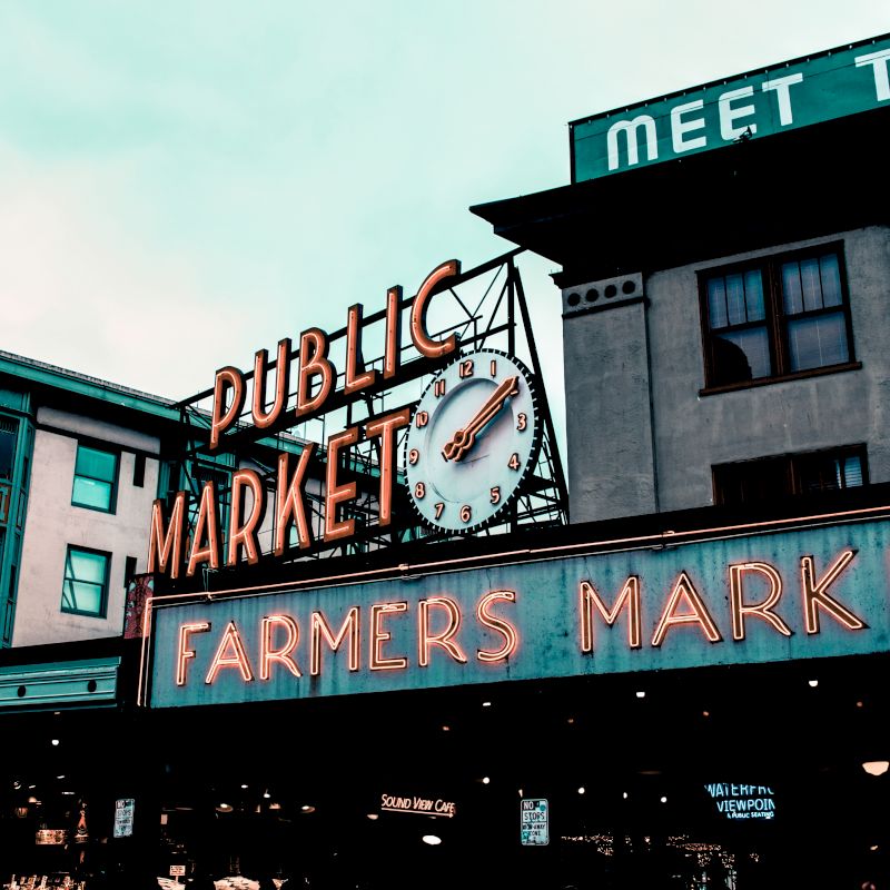 The image shows a neon sign for a farmers market, with a clock and the text "Public Market" on a building, possibly an urban setting.