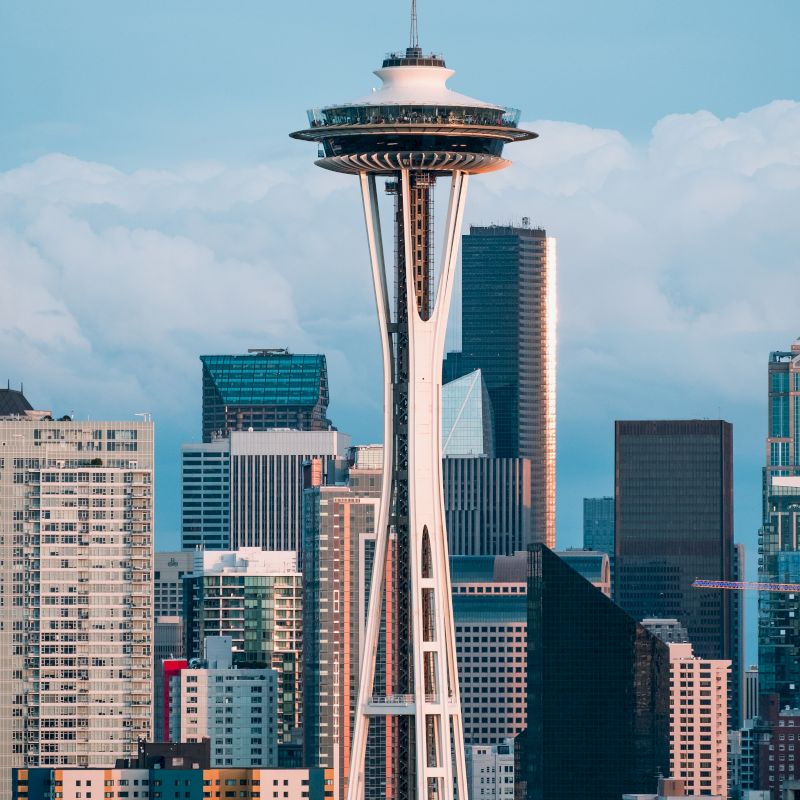 The image shows the Space Needle in Seattle, Washington, surrounded by various high-rise buildings and a partly cloudy sky in the background.