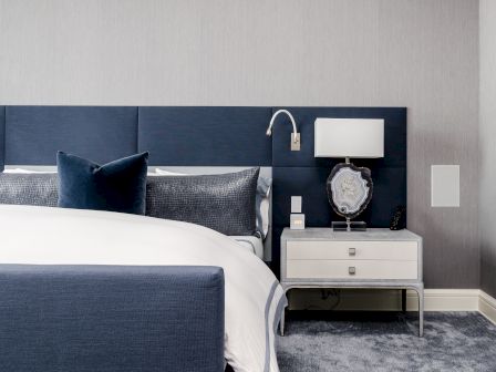 A modern bedroom featuring a blue and white theme, with a bed, plush pillows, a nightstand holding a lamp, and a decorative item.