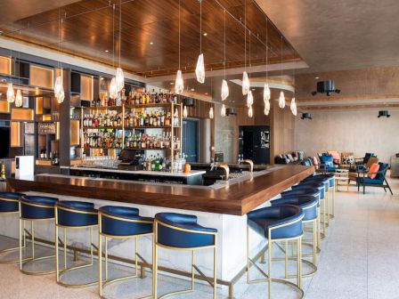 This image shows a modern bar with a wooden counter, blue chairs, a well-stocked bar shelf, and contemporary lighting fixtures.