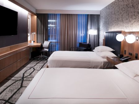 This image shows a modern hotel room with two neatly made beds, nightstands, a TV on the wall, a desk, and a chair placed by large windows.