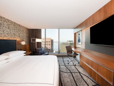 A modern hotel room with a large bed, TV, desk, chair, and a view of city buildings through a large window, ending the sentence.