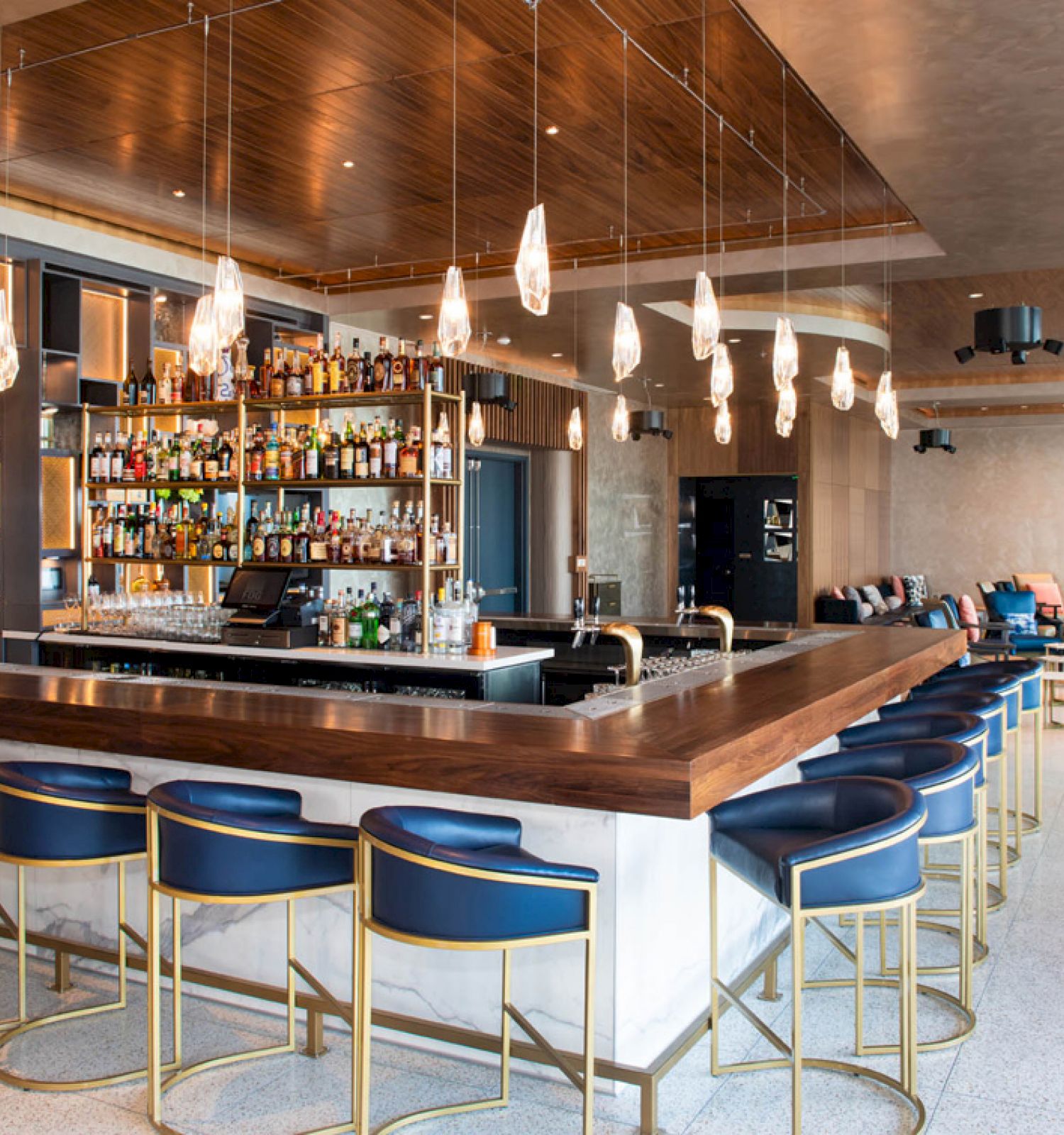 A modern bar with blue and gold stools, a well-stocked shelf, pendant lights, and a lounge area with seating in the background.
