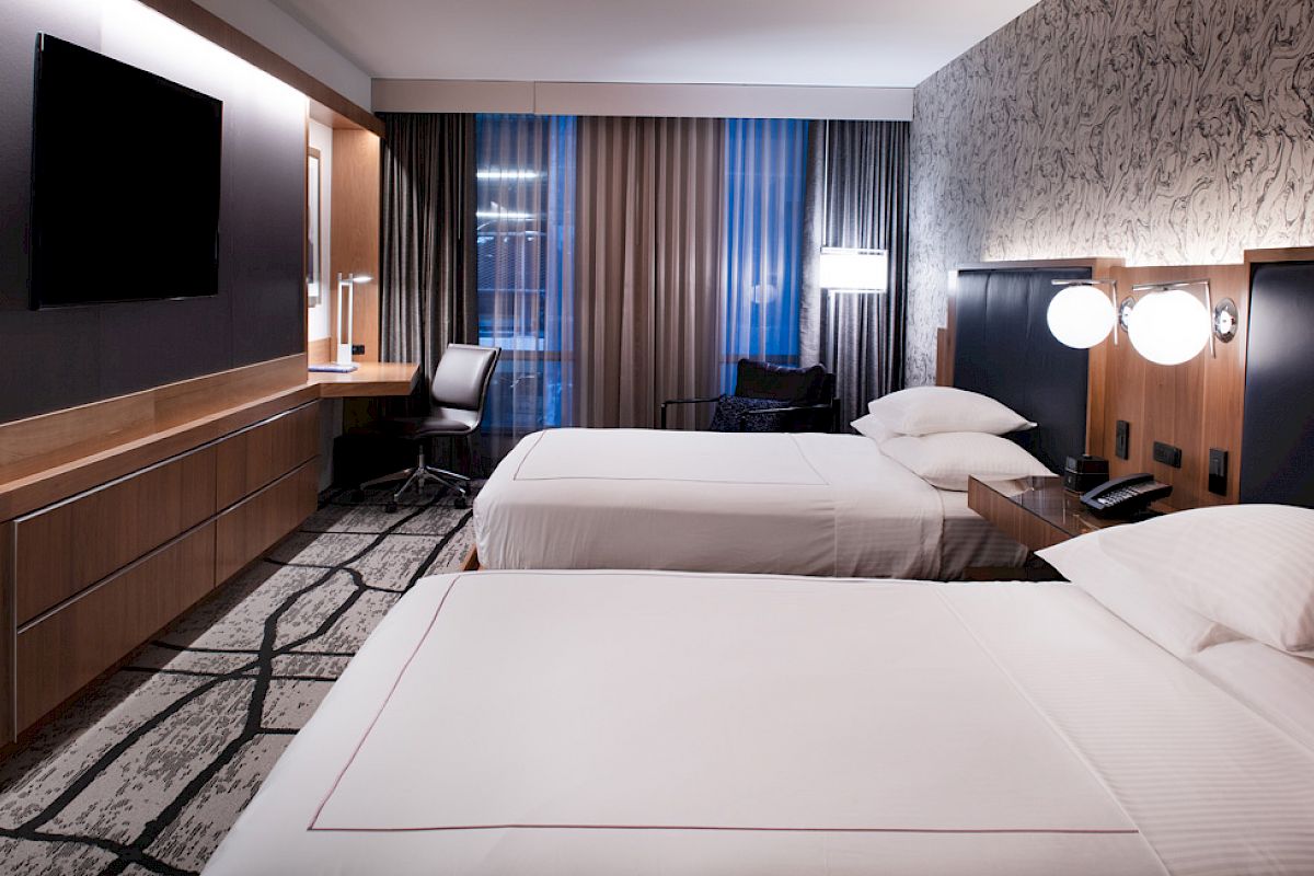 The image shows a modern hotel room with two neatly made beds, a TV, a work desk and chair, and wall lamps. The decor is contemporary and clean.