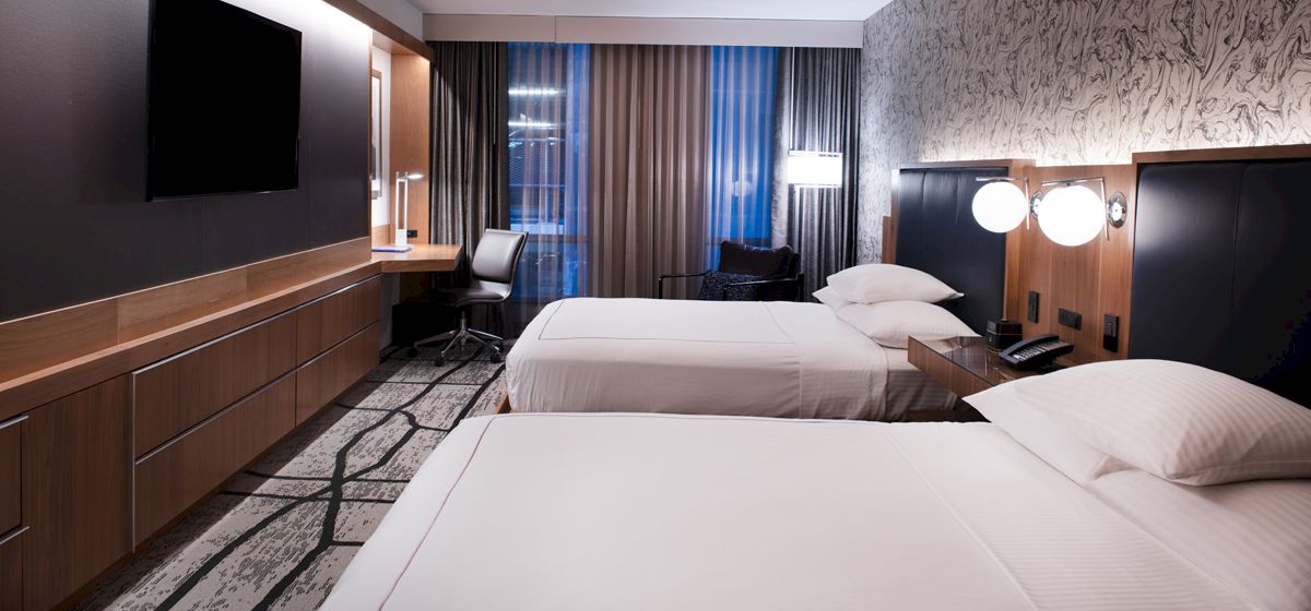 The image shows a modern hotel room with two neatly made beds, a TV, a work desk and chair, and wall lamps. The decor is contemporary and clean.