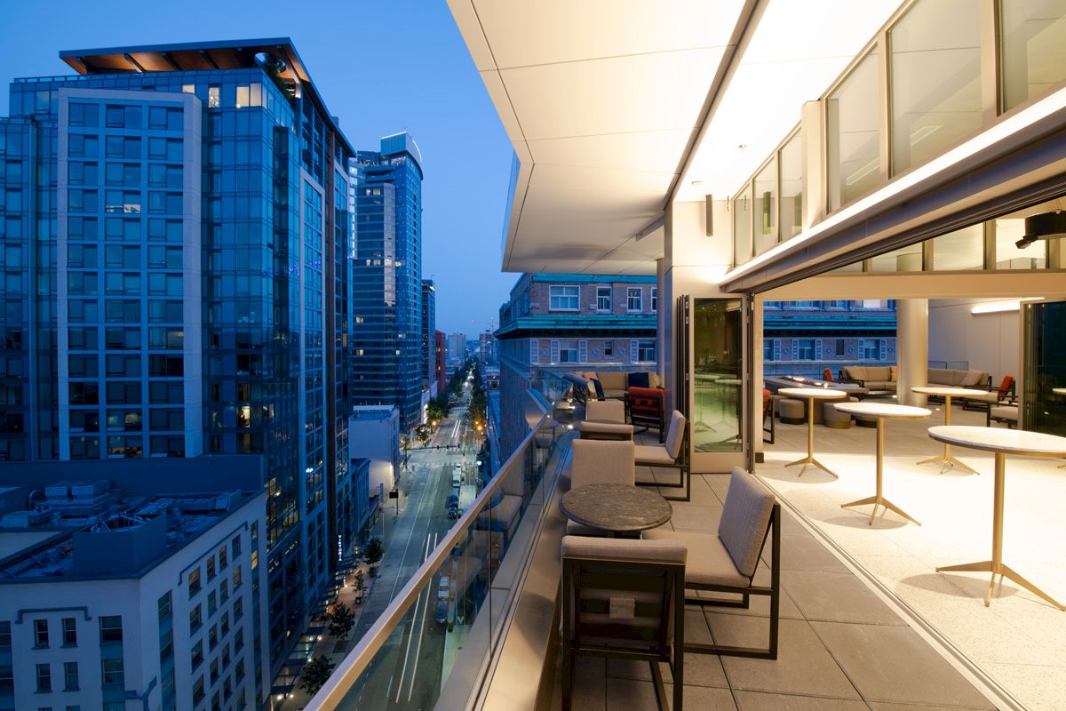 The image shows a modern outdoor balcony with tables and chairs, overlooking a city street lined with tall buildings in the evening.
