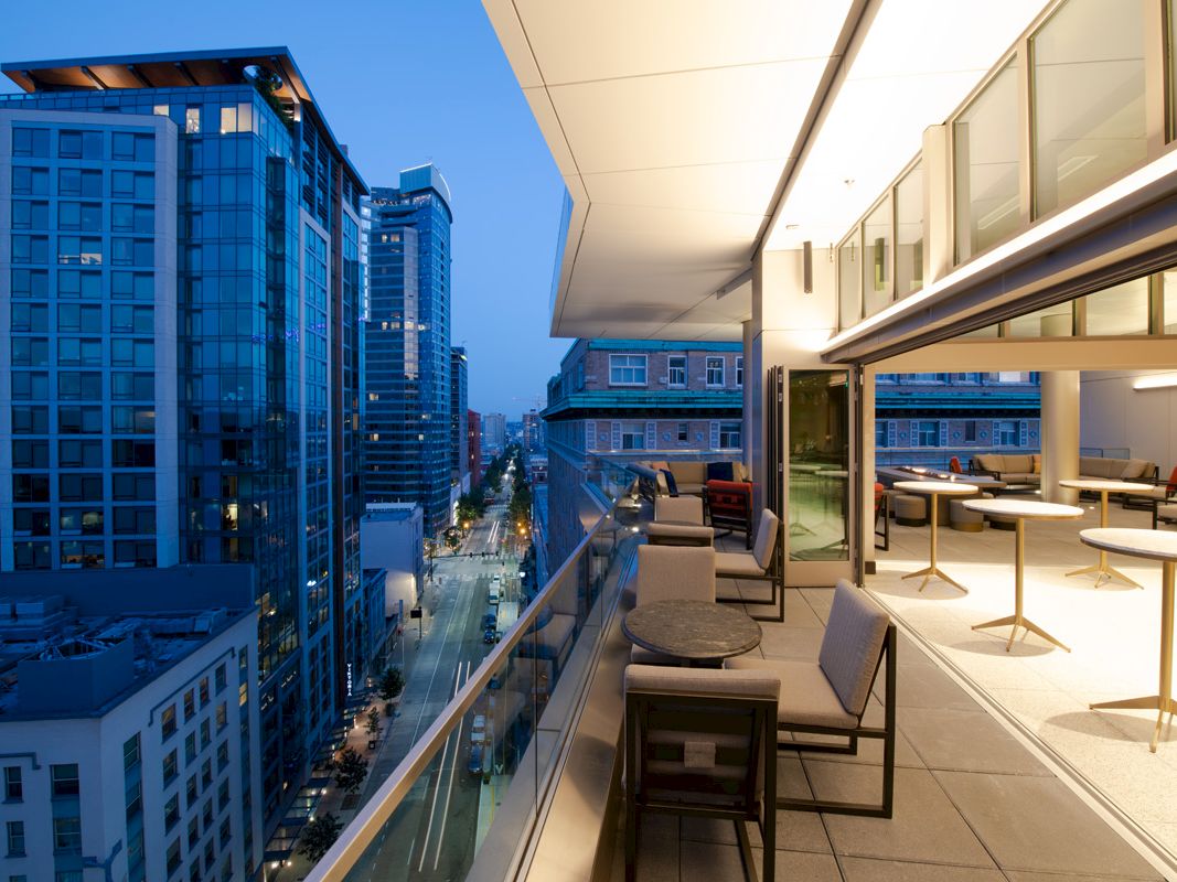 The image shows a modern outdoor balcony with tables and chairs, overlooking a city street lined with tall buildings in the evening.