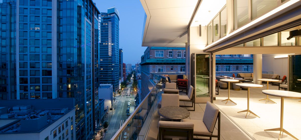 The image shows a modern outdoor balcony with tables and chairs, overlooking a city street lined with tall buildings in the evening.