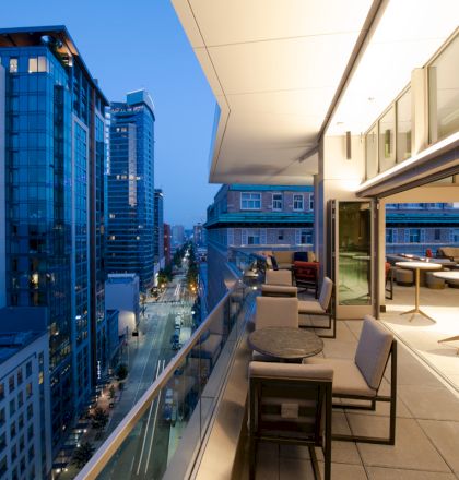 A modern, open balcony with seating and tables overlooking a cityscape at dusk, featuring tall buildings and illuminated streets below.