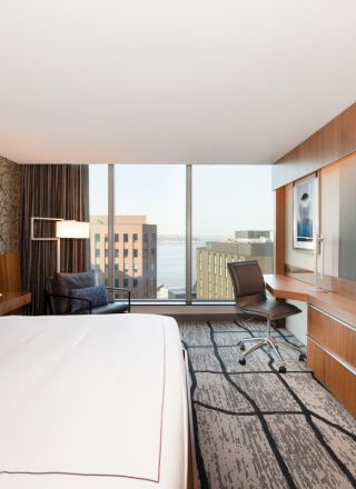 A modern hotel room with a large bed, a wall-mounted TV, a desk, a chair, and a window offering a city view.