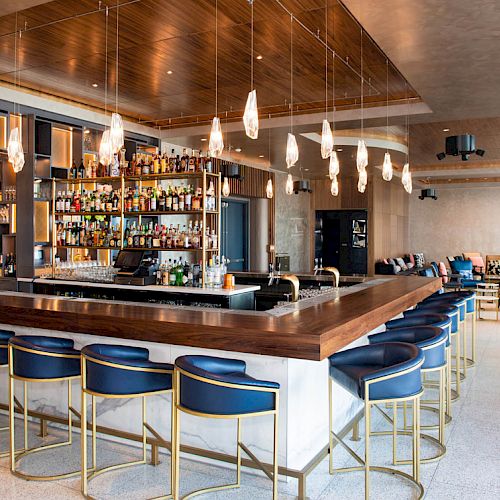 A stylish, modern bar with a central counter, surrounded by bar stools and stocked shelves of bottles in a well-lit, spacious setting.