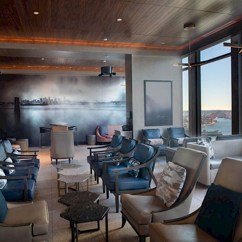 The image shows a modern lounge area with leather chairs, a mural of a cityscape, large windows offering a view of a waterfront and a ferris wheel.