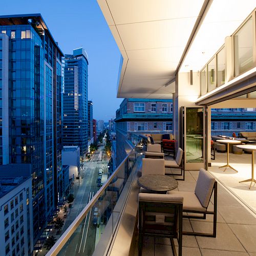A modern balcony view overlooking an urban cityscape at dusk, with tables and chairs arranged neatly along the glass railing.
