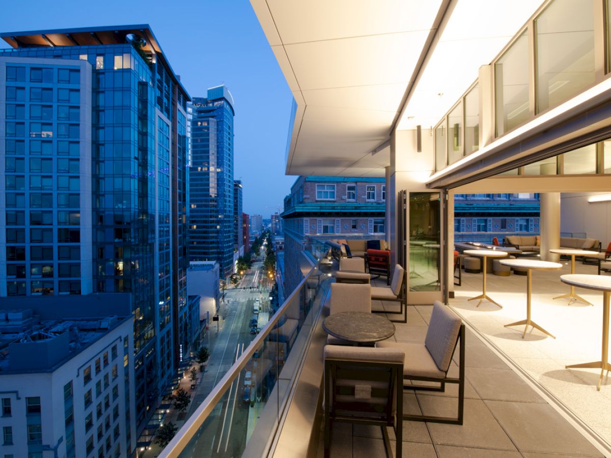 The image shows a modern balcony with tables and chairs overlooking a city street with tall buildings in the evening.