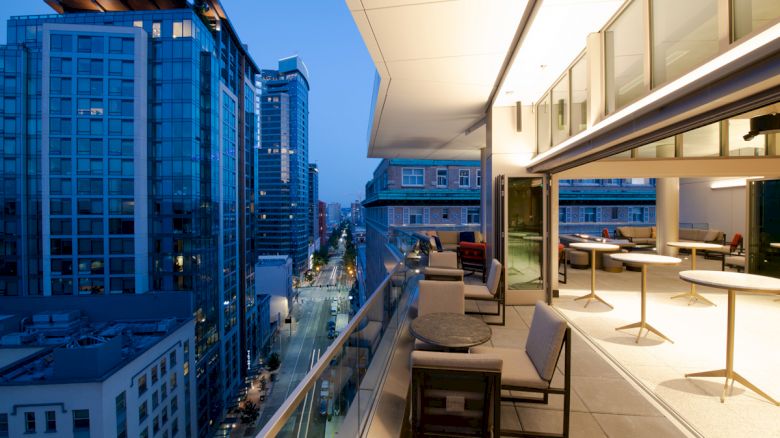 The image shows a modern balcony with tables and chairs overlooking a city street with tall buildings in the evening.