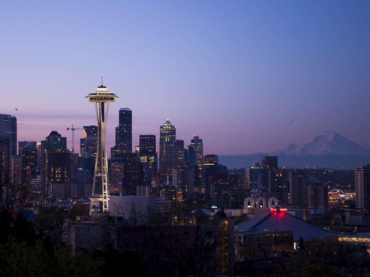 The image shows a cityscape at dusk with the Space Needle, skyscrapers, and mountains in the background against a twilight sky.