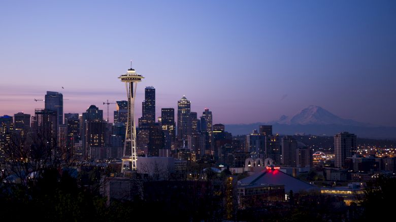 The image shows a cityscape at dusk with the Space Needle, skyscrapers, and mountains in the background against a twilight sky.
