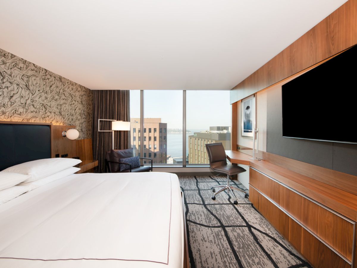 A modern hotel room features a large bed, a desk with a chair, flat-screen TV, and a window with cityscape and waterfront views.