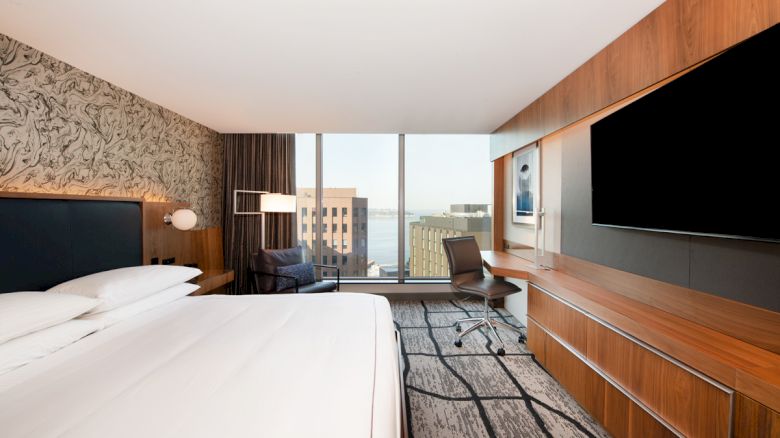 A modern hotel room features a large bed, a desk with a chair, flat-screen TV, and a window with cityscape and waterfront views.