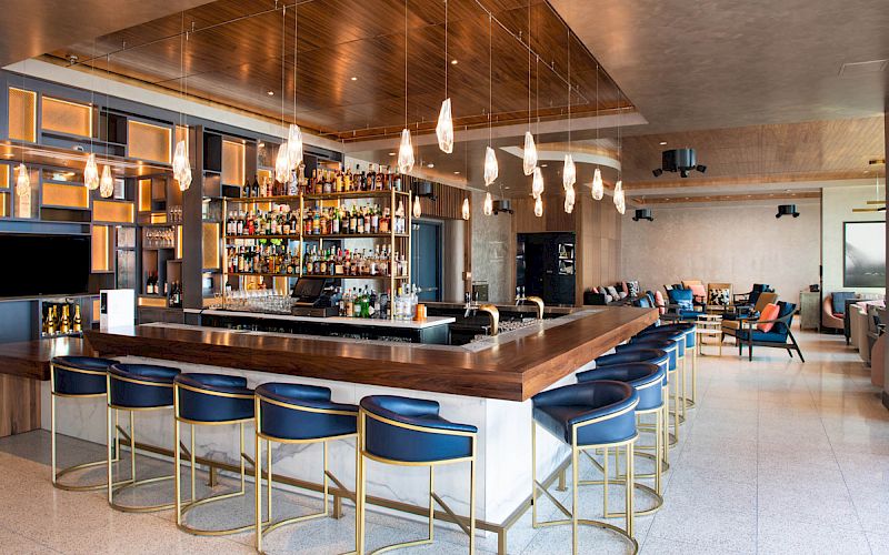 This image shows a modern bar area with a row of blue barstools, hanging pendant lights, and a stocked bar. The space has a contemporary design.
