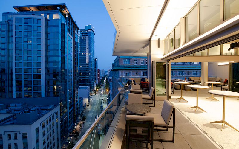 A modern high-rise balcony with outdoor seating overlooks a city street at dusk, with tall buildings visible in the background.