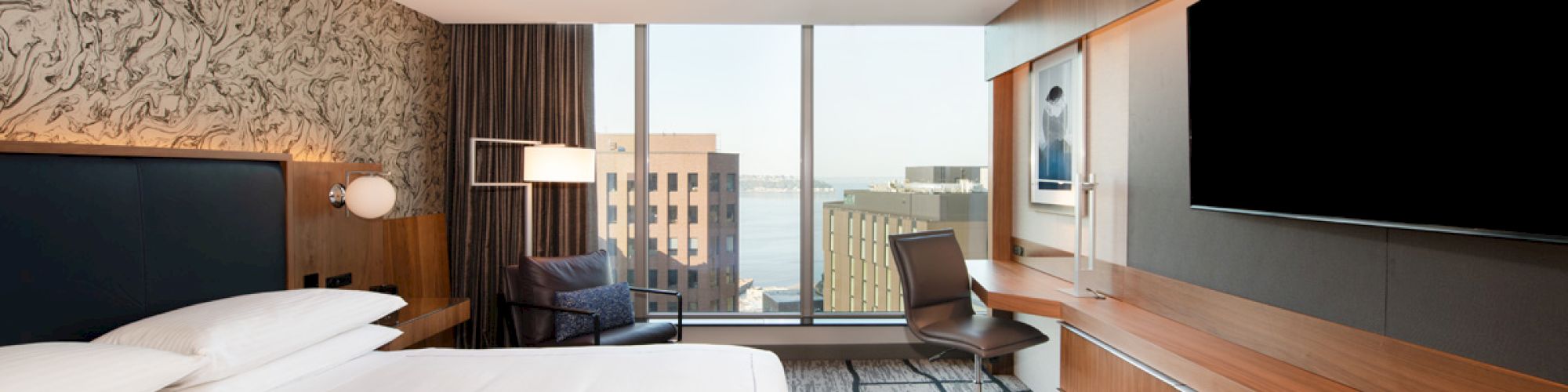 A modern hotel room with a king-sized bed, large TV, desk, chair, city view through large windows, and contemporary decor finishes the sentence.